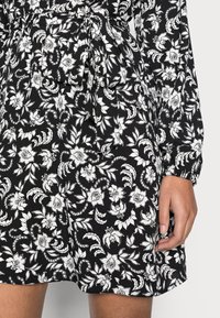 Vestido de tecido preto com um padrão floral branco, mangas longas, cintura ajustada com um laço e punhos franzidos nas mangas.