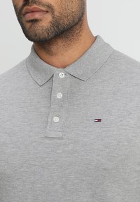 Grijze polo t-shirt van textuurstof, met een drieknopensteek en een klein geborduurd logo in blauw, rood en wit.