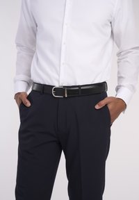 Schwarzer Ledergürtel mit einer silbernen Metallschnalle, getragen über einer navyblauen Hose und einem weißen Hemd mit Knöpfen. Glatte Textur, klassisches Design.