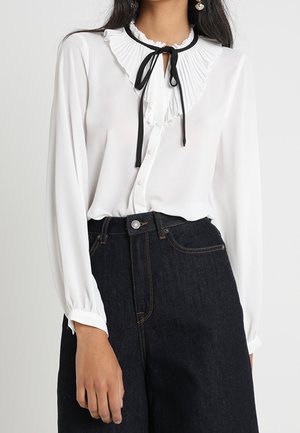 Femme portant un chemisier blanc à manches longues avec col plissé et ruban noir noué, associé à un jean en denim foncé taille haute.