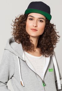 Sudadera gris con cremallera y cordones blancos, llevada sobre una camiseta blanca. Gorra negra con visera verde. Cabello rizado visible.