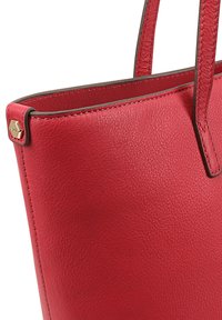 Sac fourre-tout en cuir rouge avec une texture lisse, doté de deux poignées et d'un accent hexagonal en or. Aucune fermeture ou motif visible.