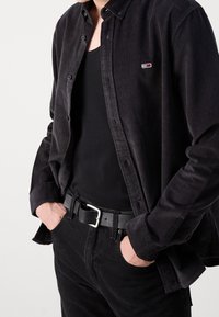 Camisa de pana negra con cierre de botones, llevada sobre una camiseta sin mangas negra. Combinada con pantalones negros y un cinturón negro con una hebilla plateada.