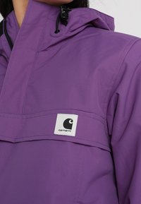 Lilla vandtæt jakke med høj krave, justerbar hætte og en hvid firkantet logo på brystområdet. Glat tekstur og holdbart materiale.