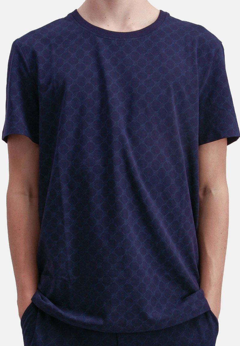 Man in een donkerblauw T-shirt en bijpassende broek met subtiele geometrische patronen, met zijn handen in zijn zakken, tegen een effen achtergrond.