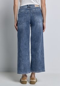 Jeans larges bleu clair en denim, présentant un dos épuré avec deux poches et une taille simple, associés à des baskets beiges et blanches.