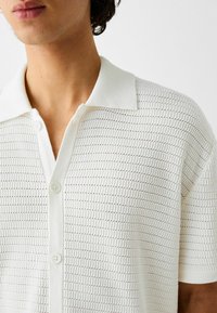 Camiseta polo blanca de manga corta confeccionada en tejido de punto texturizado, con cuello clásico y tapeta de tres botones con sutiles rayas horizontales.