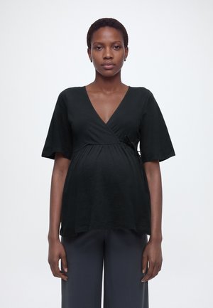 Rase naine, kes kannab musta lühikeste varrukatega wrap-tüüpi topi ja tumedaid pükse, seisab ühevärvilise valge tausta ees.