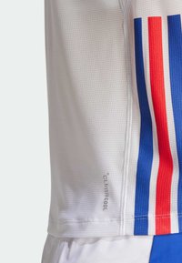 La camiseta atlética blanca presenta franjas rojas y azules a los lados. Se puede ver el texto "climacool". La tela tiene un acabado texturizado.