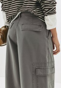 Pantaloni cargo grigi a gamba larga con due tasche applicate sui lati, visti da dietro, abbinati a una maglietta a maniche lunghe a righe e a una borsa marrone.