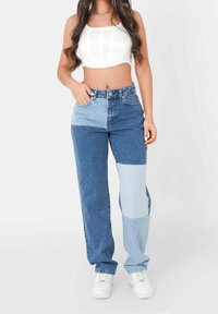 Mujer vistiendo unos vaqueros de parchís en dos tonos de azul, una camiseta blanca corta y zapatillas blancas, de pie con una mano en el bolsillo.