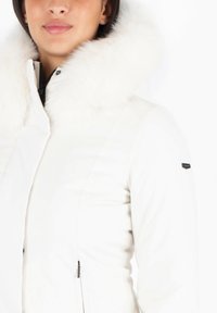 Chaqueta aislante blanca con un grueso cuello de piel sintética, que cuenta con un cierre de cremallera y un pequeño detalle de marca en la manga izquierda.