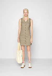 Vestido cáqui com botões, decote em V, design sem mangas e bolsos frontais. Combinado com uma mala tote creme e ténis brancos.