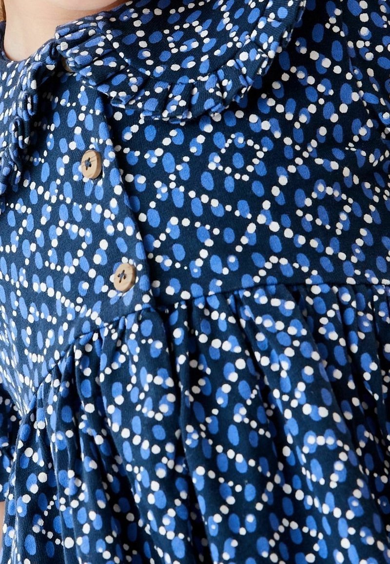 Robe bleue avec un motif à pois blancs et bleu clair, avec un col à volants et des boutons marron sur le devant.