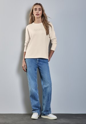 Beige overhemd met lange mouwen en een reliëf "ADORN" logo, gecombineerd met lichtblauwe rechtlopende jeans en witte sneakers, staande tegen een lichte achtergrond.