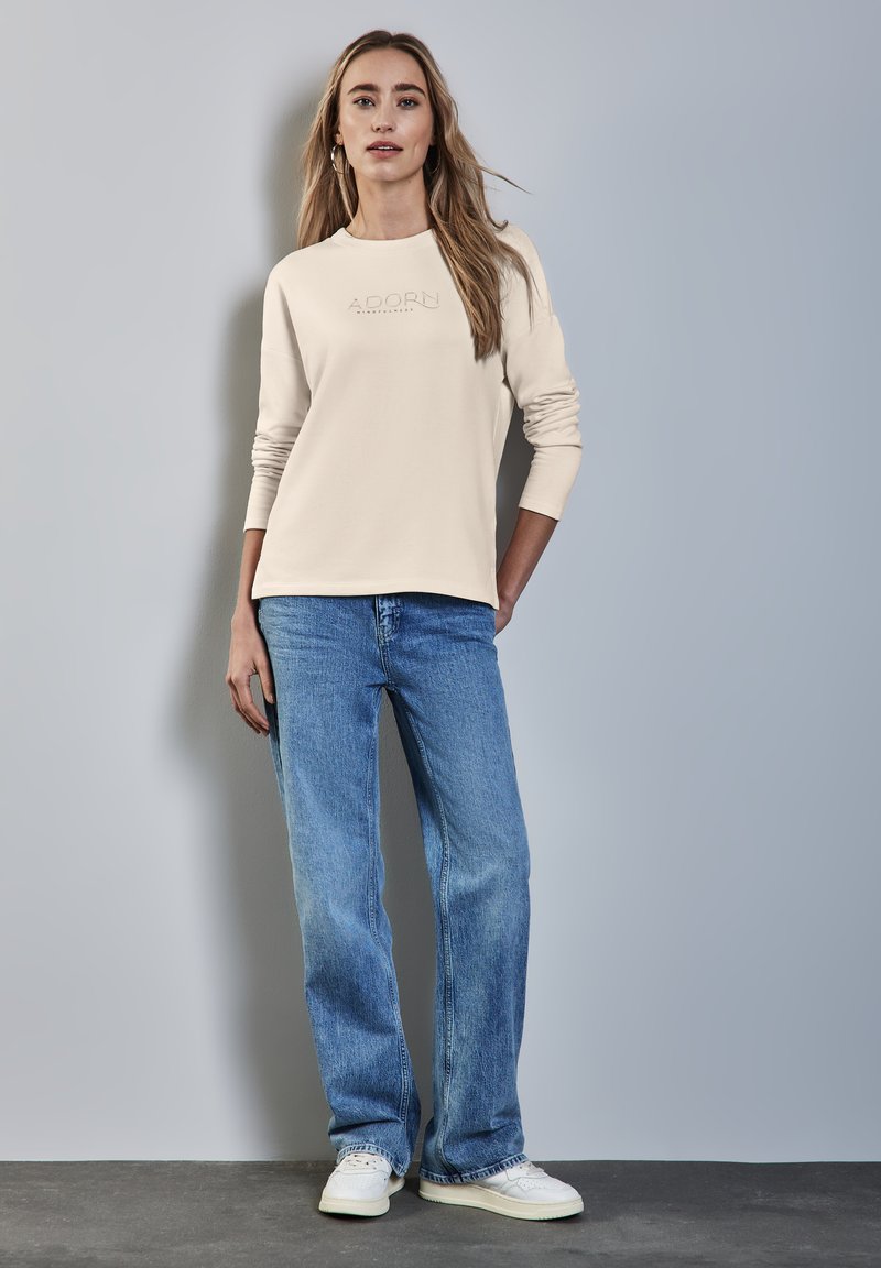 Beige Langarmshirt mit eingeprägtem "ADORN"-Logo, kombiniert mit hellblauen gerade geschnittenen Jeans und weißen Sneakers, stehend vor einem hellen Hintergrund.