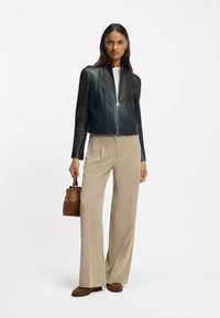 Zwarte leren cropped jas met een hoge kraag, gecombineerd met beige wijde broekspijpen. Het model houdt een kleine bruine suède handtas vast.