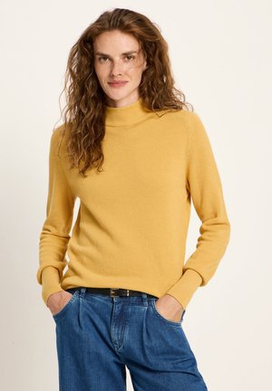 Woman with long curly hair wearing a mustard yellow sweater tucked into blue jeans with hands in pockets, standing against a plain background.