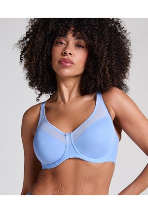 Vrouw met krullend haar die een lichtblauwe beugelbeha met mesh-details draagt, zelfverzekerd poseert tegen een effen grijze achtergrond.