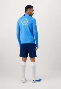 Blå langærmet trænings T-shirt med gul "THFC" logo på ryggen, parret med mørkeblå shorts og hvide sokker, iført blå fodboldsko.