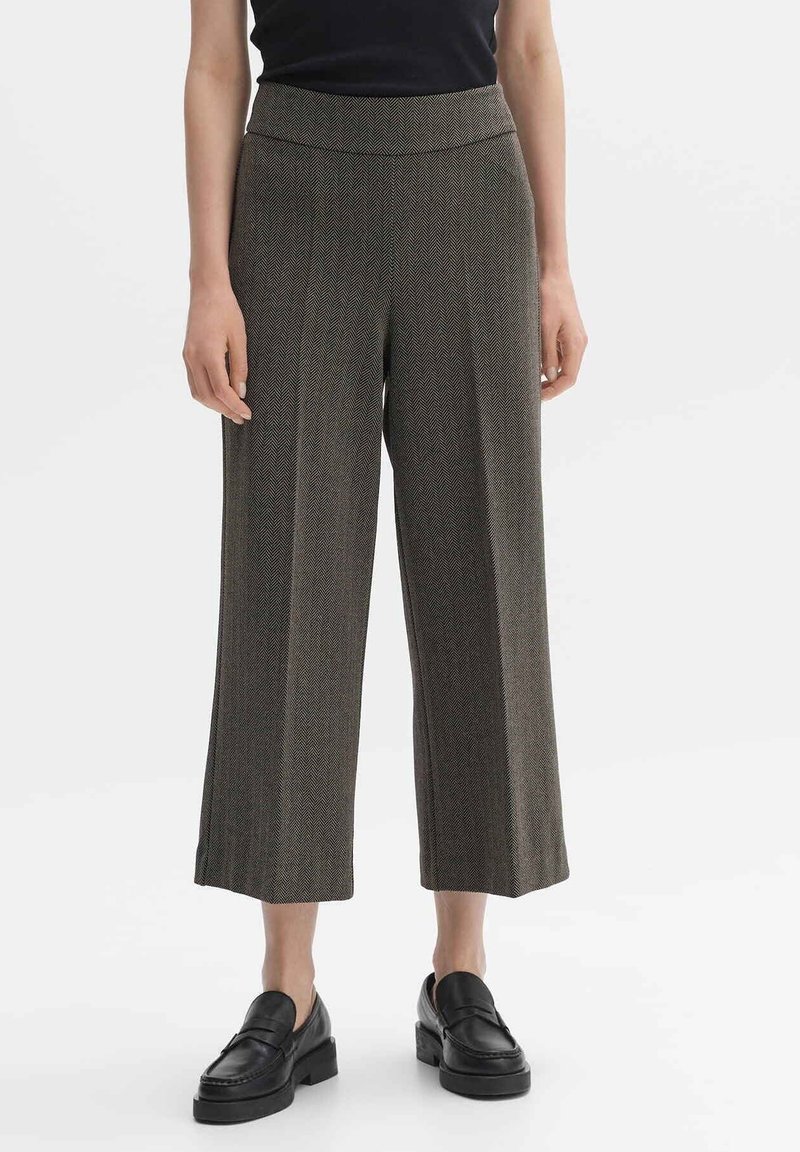 Hnědé široké kalhoty s herringbone vzorem a vysokým pasem. V kombinaci s černými mokasíny, které mají hladkou texturu a upravený design.