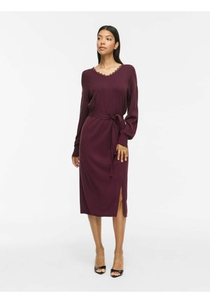 Femme portant une robe en maille bordeaux à manches longues avec ceinture à la taille et fente latérale, associée à des talons ouverts noirs.