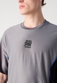 Pilkas trumpas rankovės sportinis marškinėliai su tekstūruotu audiniu, apvalia iškirpte, juodu logotipu krūtinėje ir mėlynu akcentu šone.