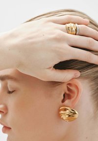 Pendientes de oro con un diseño ondulado, combinados con un anillo texturizado en oro y plata. Las superficies suaves reflejan la luz, realzando su forma.