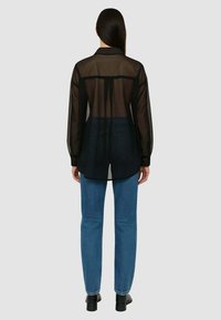 Camicia nera trasparente a maniche lunghe con schiena semitrasparente, abbinata a jeans scuri. Presenta un colletto e un taglio dritto.