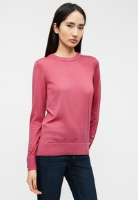 Ροζ πουλόβερ με μακριά μανίκια από απαλό υλικό, με στρογγυλή λαιμόκοψη, πλαϊνές σχισμές και κουμπιά στις μανσέτες. Συνδυασμένο με σκούρο τζιν.