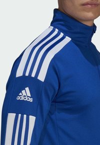 Blauwe sportjas met een hoge kraag, drie witte strepen op elke schouder en een geborduurd wit Adidas-logo op de mouw.