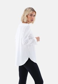 Camicia bianca a maniche lunghe con orlo arrotondato, polsini con bottone e una morbida texture. Indossata con pantaloni scuri, creando un contrasto di colore.