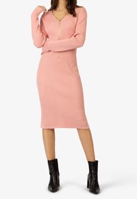 Femme portant une robe midi rose côtelée à manches longues avec une fermeture éclair frontale et des bottines noires, debout les bras croisés.