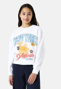 Sudadera blanca con texto azul "NEW YORK" y gráfico colorido de frutas. Combinada con pantalones navy a rayas. Material de algodón, corte relajado.