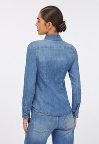 Cămașă din denim de culoare albastră deschisă, cu un design ajustat, mâneci lungi, manșete cu nasturi și guler. Afișată din spate, evidențiind detaliile cusăturii.