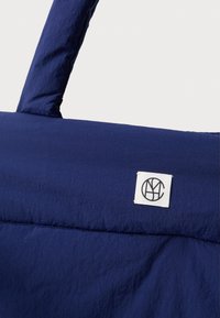 Tmavě modrá látková taška s polstrovanými rukojetmi a malou bílou čtvercovou nálepkou se šitým černým kruhovým logem na přední straně.