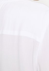 Camicia bianca realizzata in un tessuto semi-trasparente, con una texture liscia e una cucitura orizzontale lungo la parte superiore della schiena.