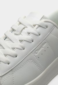 Λευκό δερμάτινο αθλητικό παπούτσι με υφαντές λευκές κορδόνια, ορατές ραφές και διακριτική ανάγλυφη σήμανση στην πλευρική επιφάνεια.