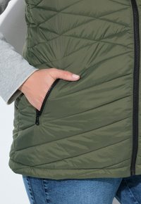 Olivgrüne, gestepptes West, mit diagonaler Steppung und einer Reißverschlusstasche. Hellgraues Langarmshirt ist darunter sichtbar.