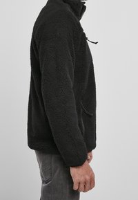Schwarze Fleecejacke mit hohem Kragen, Reißverschluss vorne und Seitentaschen. Strukturmaterial mit einem weichen, flauschigen Aussehen.