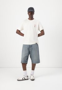 Tricou alb cu mânecă scurtă și grafică roz, șorturi din denim albastru și adidași albi cu accente negre. Modelul poartă o șapcă închisă la culoare.