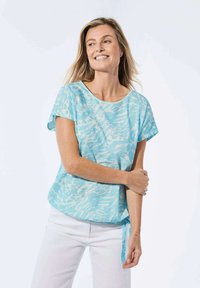 Helles Blaues Kurzarmoberteil mit strukturiertem Muster, runder Ausschnitt und seitlichem Bindeband. Getragen mit weißen Hosen, vor einem neutralen Hintergrund.