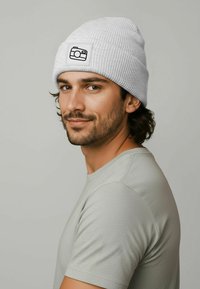 Gray ribbed beanie with a black camera graphic on a patch, worn by a person in a light gray t-shirt, with medium-length dark hair.