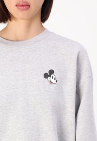Világosszürke pulóver, amelyen a bal mellrészen egy kis fekete-fehér Mickey Mouse-logó található. Puha, texturált anyag.