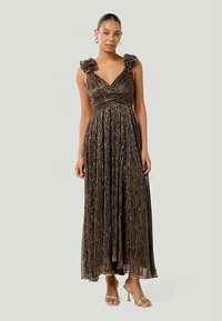 Femme portant une robe de soirée sans manches en métal plissé avec des bretelles à volants et des sandales à talons hauts dorées.