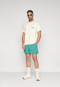 Μπεζ βαμβακερό μπλουζάκι με τσέπη στο μπροστινό μέρος, συνδυασμένο με πετρόλ σορτς. Συμπληρώνεται με μπεζ γυαλιά ηλίου και λευκές κάλτσες, φορώντας αθλητικά παπούτσια.