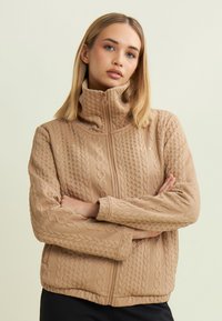 Femme aux cheveux blonds portant une veste matelassée beige à zip avec des motifs de tricot en relief, se tenant les bras croisés devant un fond uni.