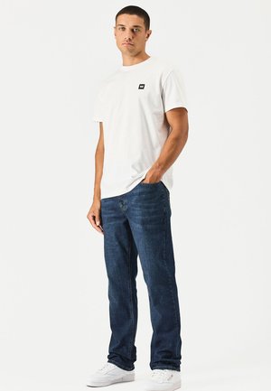 Junger Mann, der mit einer Hand in der Tasche steht, trägt ein weißes T-Shirt, blaue Jeans und weiße Sneakers vor einem einfarbigen Hintergrund.