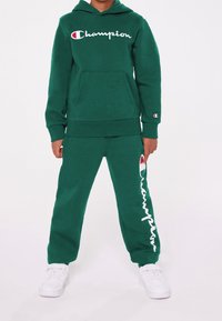Grön bomullshoodie med vit "Champion" logotyp, framficka, matchande byxor med logotyp längs sidan, kombinerat med vita sneakers.