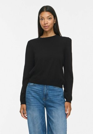 Kvinde iført en sort langærmet top med let polstrede skuldre og blå jeans, stående mod en ensfarvet hvid baggrund.